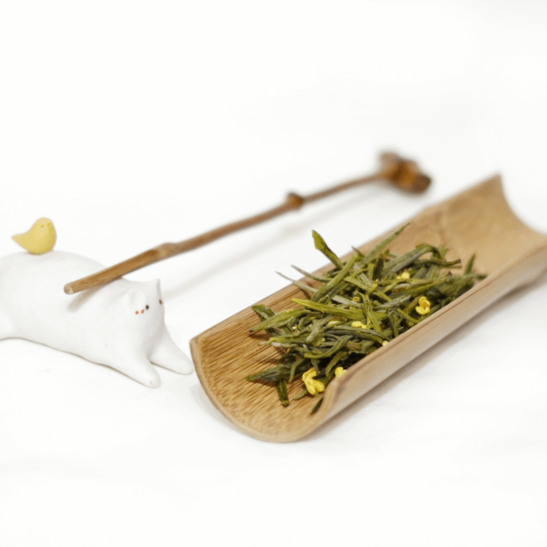 A wooden plate with green tea leaves and a touch of osmanthus flowers tea, alongside a white teapot and chopsticks on a white background.