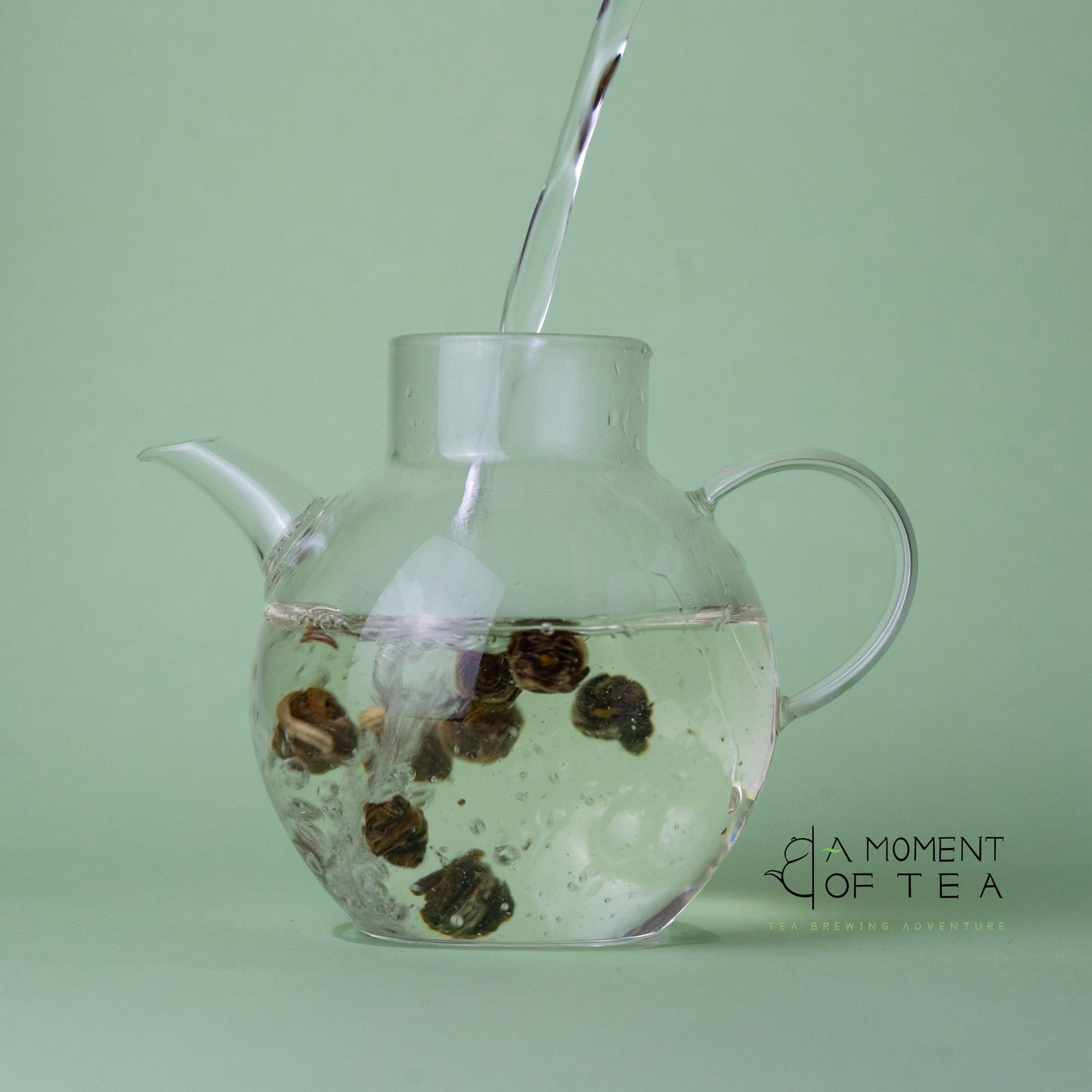 Glass teapot with Jasmine Dragon Pearls green tea being filled with water against a green background.
