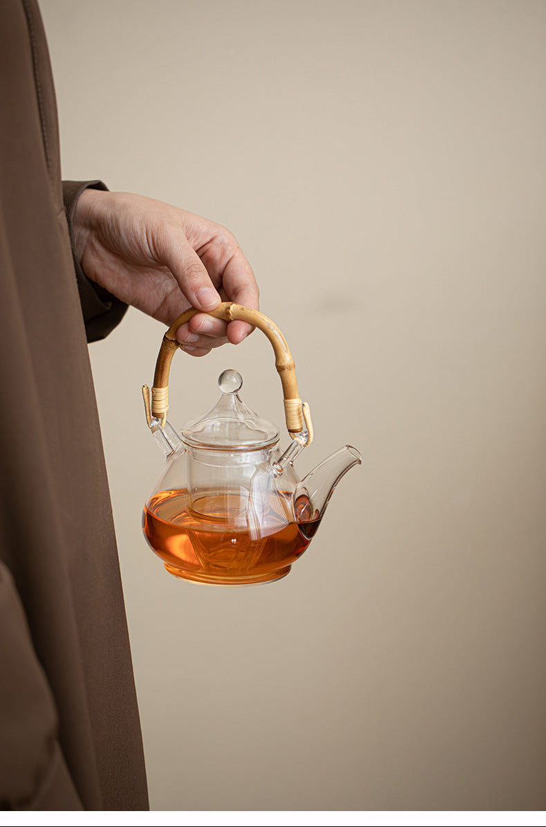 A hand holds a clear glass teapot with a bamboo handle and spout, filled with amber tea, against a warm beige background.
