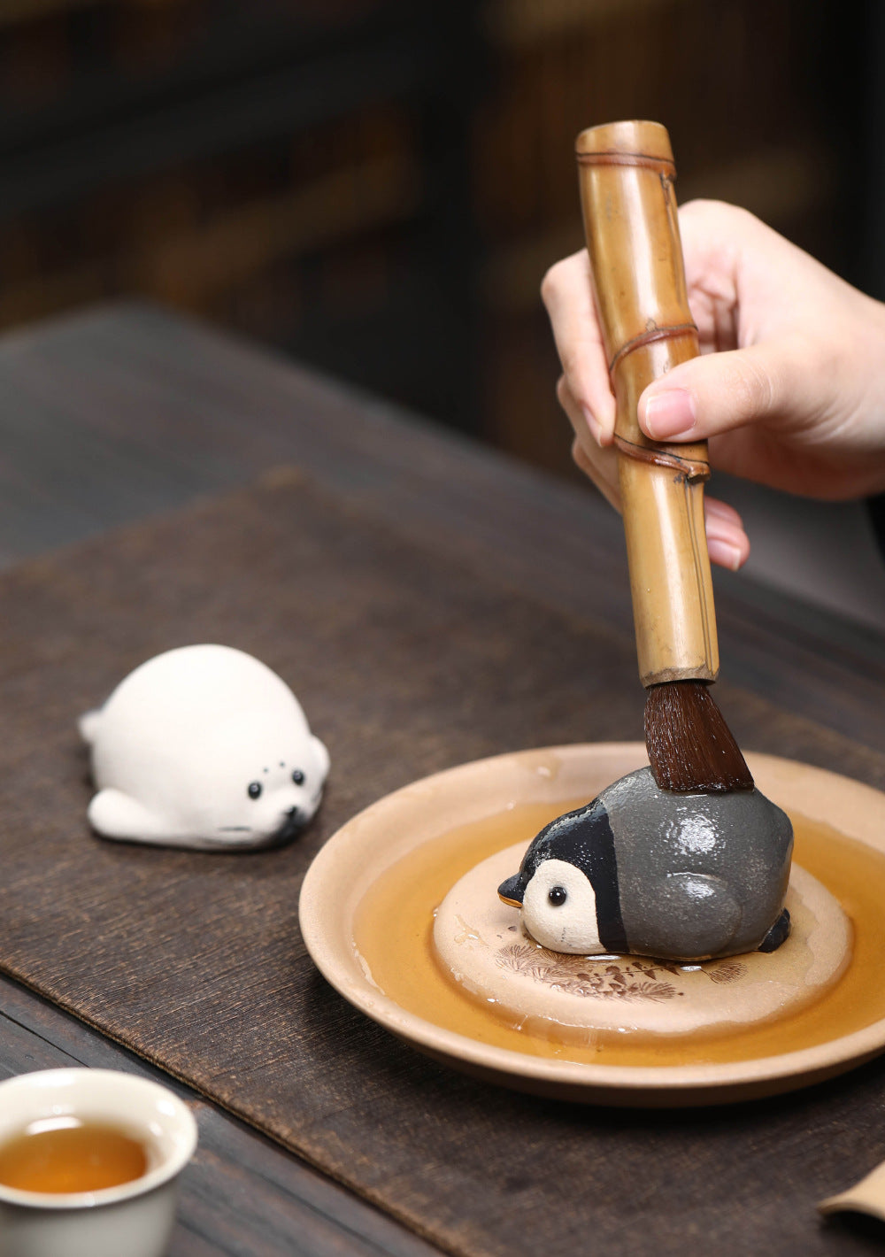 Wet penguin tea pet groomed with a bamboo tea brush on a dish, seal tea pet nearby