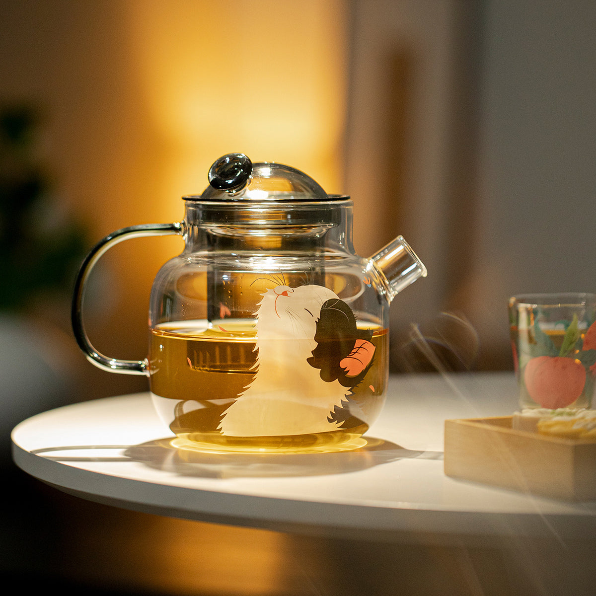 Cat and butterfly glass teapot filled with tea on a round table beside a glass cup