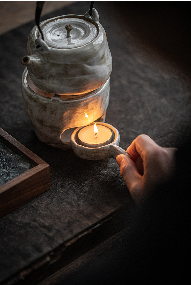 Ceramic teapot warmer with a lit candle inside on a dark surface, hand holding a small ceramic container.