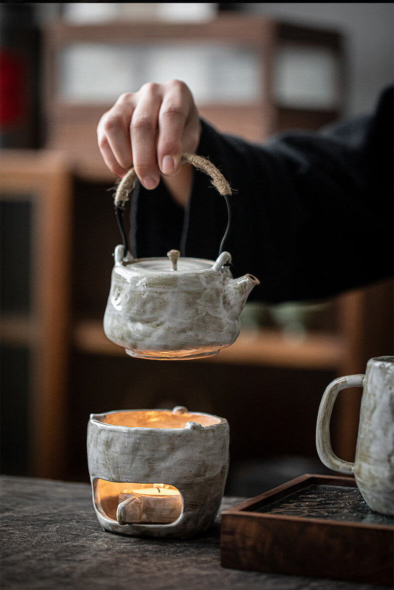 Person holding a small ceramic teapot with warmer on a wooden surface.