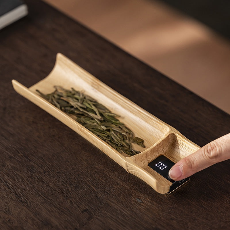 Bamboo tea scale on a dark wood table with green tea leaf in the trough and the weight showing