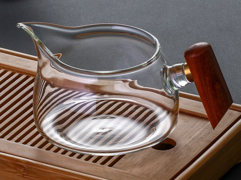 Wooden Handle Glass Fairness Cup resting on a bamboo tea tray, beak spout facing left