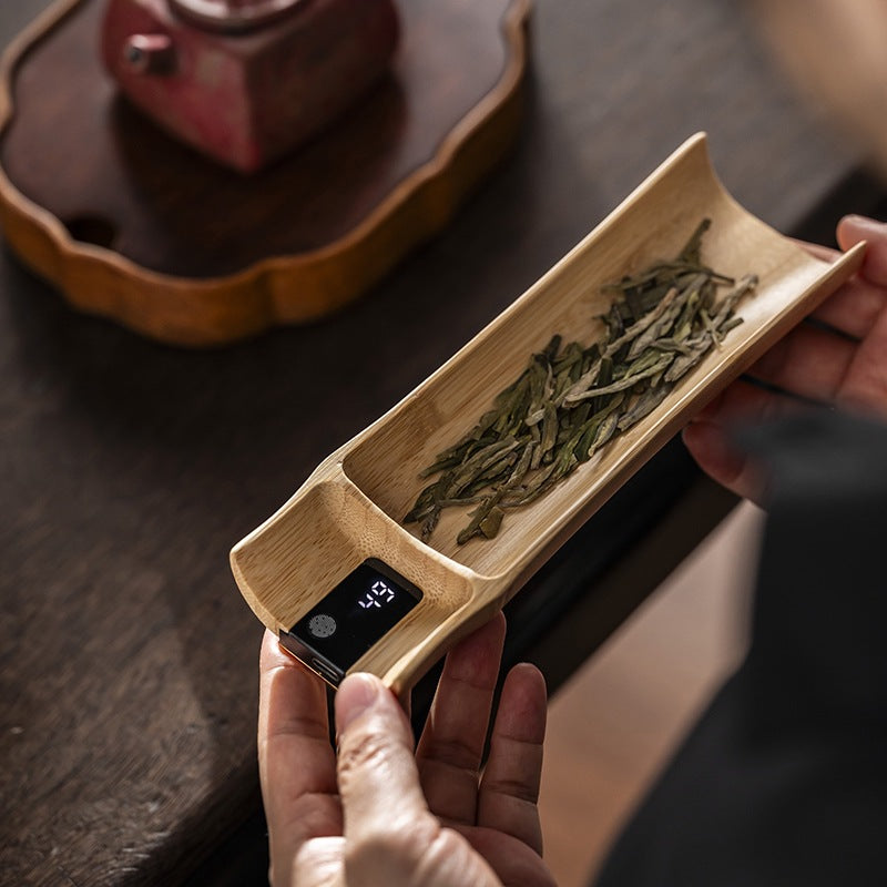 Hands holding the bamboo tea scale with loose green tea leaf, digital readout visible