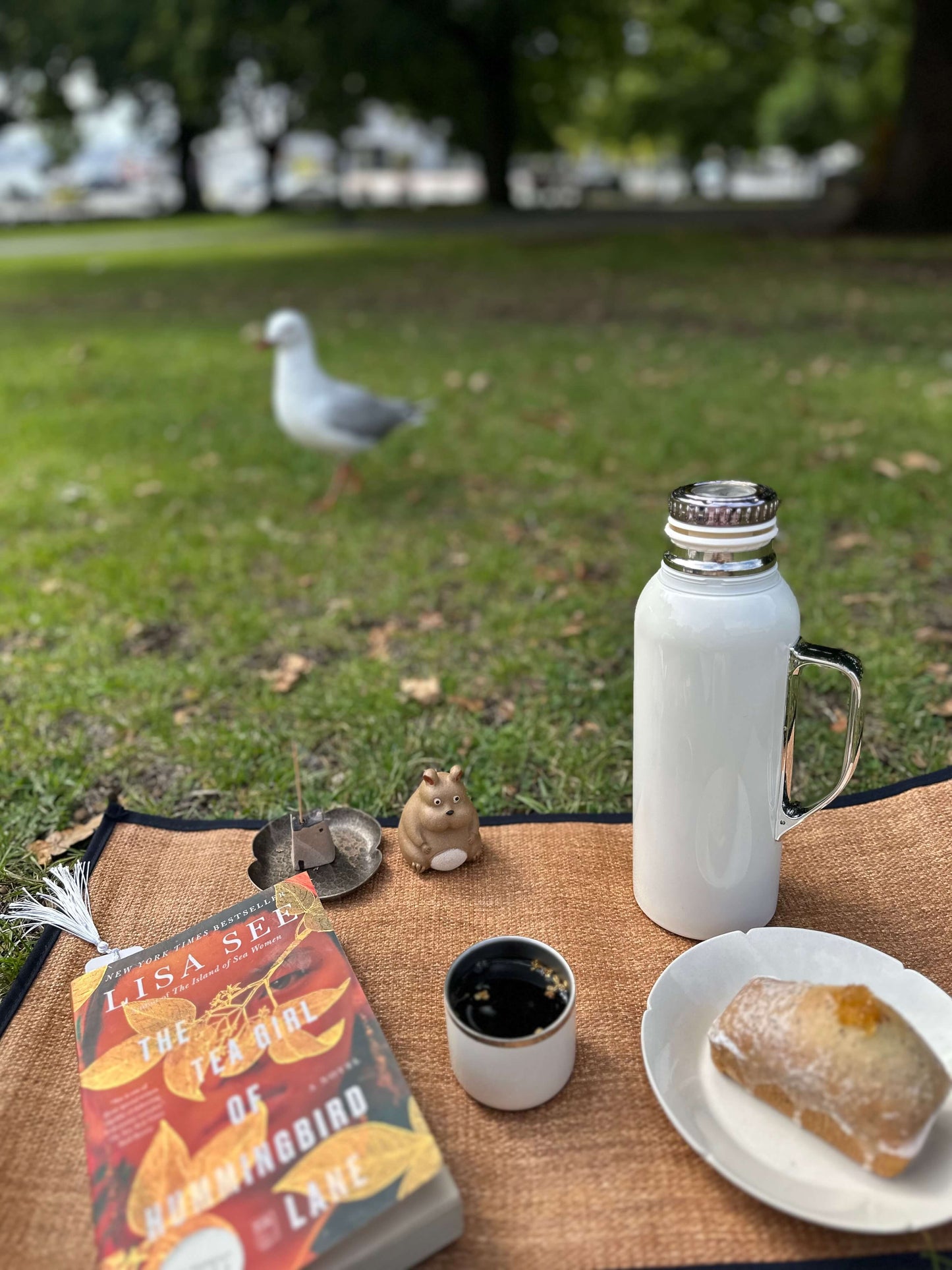 Outdoor setting with a book, coffee, pastry, and thermos on a table with a seagull in the background.