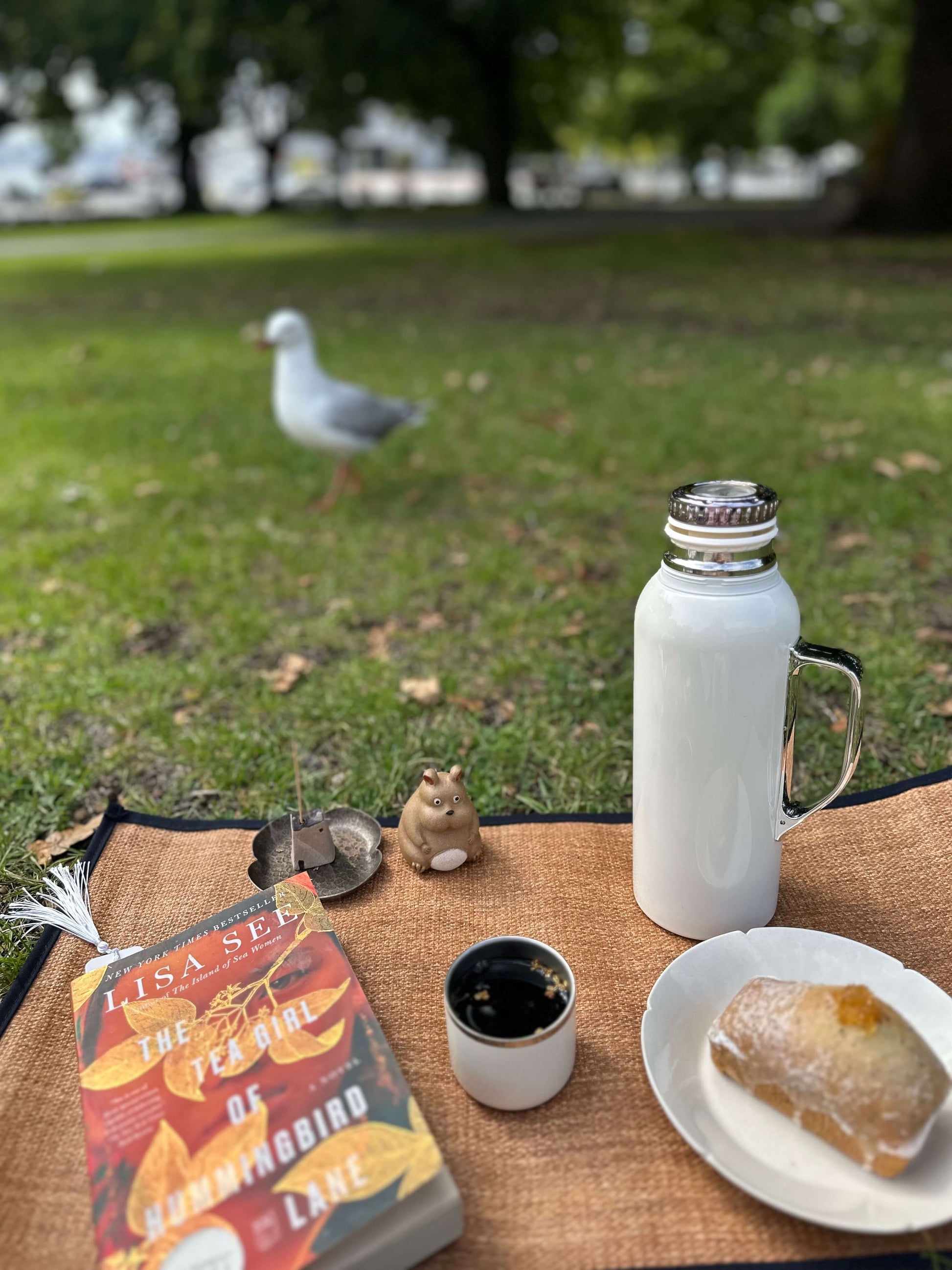 Outdoor setting with a book, coffee, pastry, and thermos on a table with a seagull in the background.