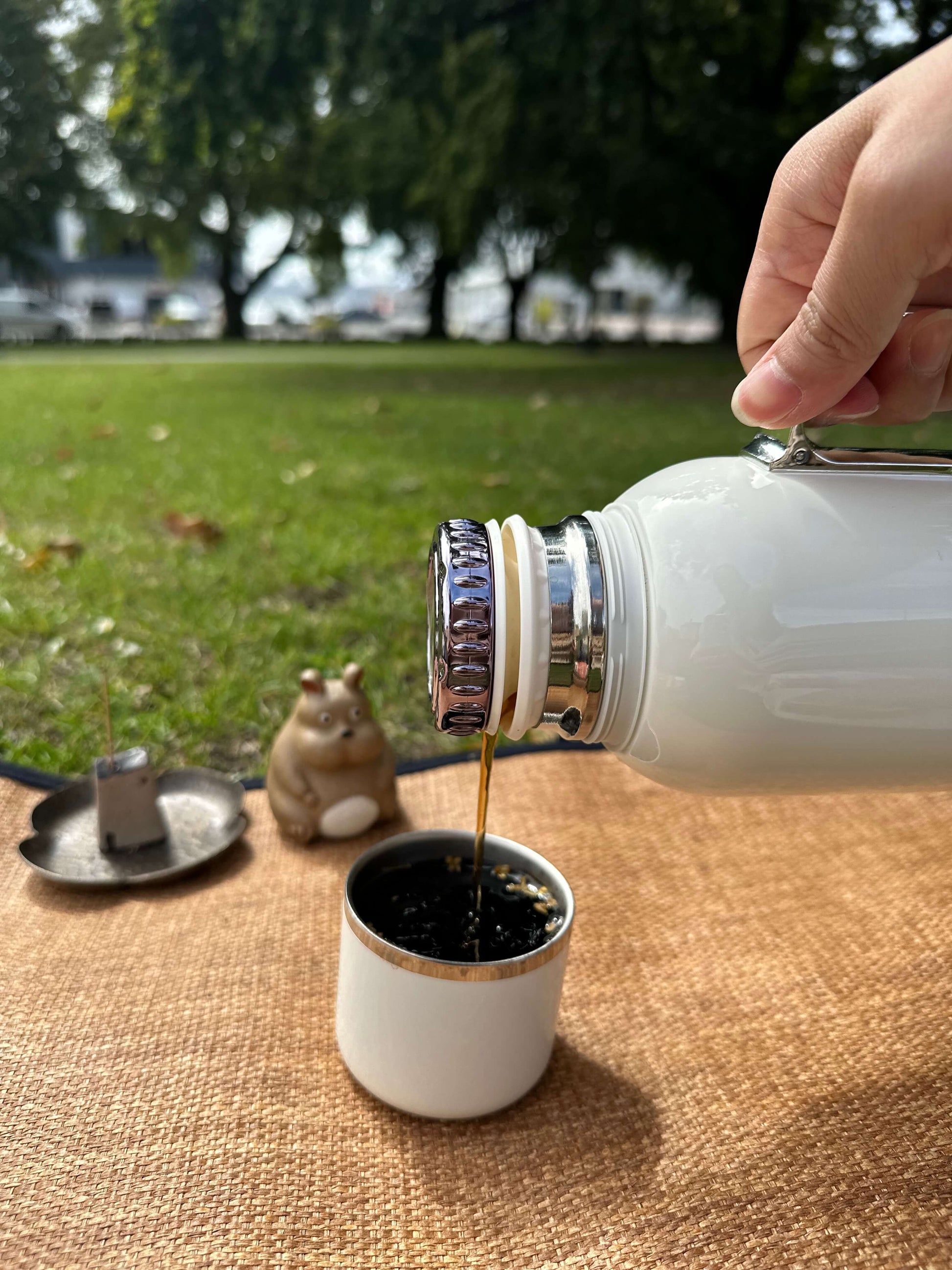 Person pouring liquid from a white tea flask into a small pot on a table outdoors.
