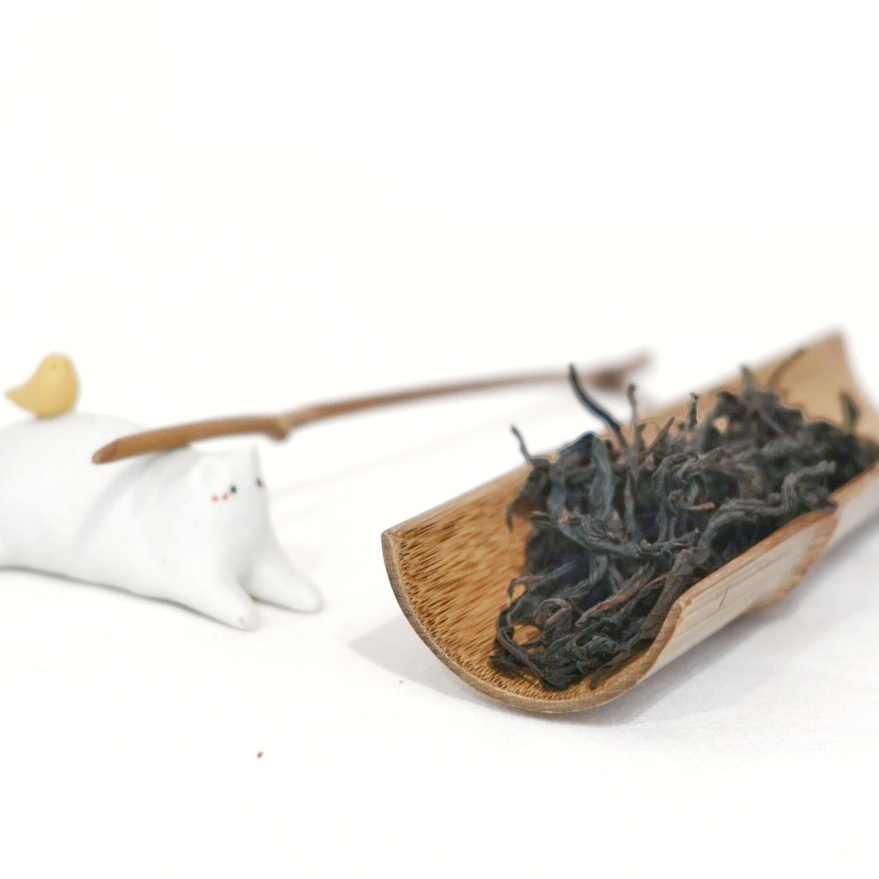 A small amount of smoked Lapsang Souchong tea leaves displayed on a wooden dish, with a white tea bag adorned with a heart shape in the background.