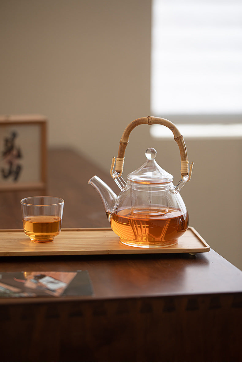 A clear glass teapot filled with amber tea sits on a bamboo tray next to a matching glass cup on a wooden table, with a softly blurred indoor background.