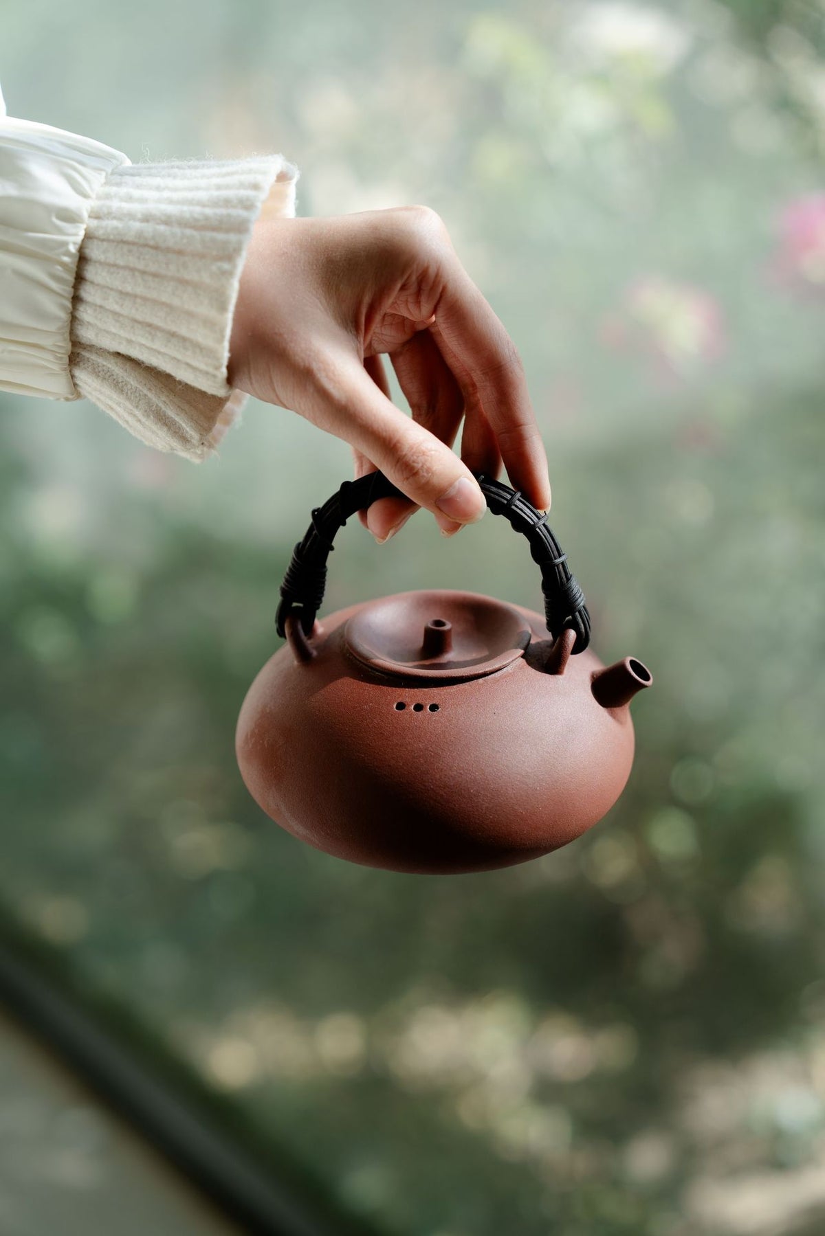 Rattan handle clay boiling teapot held in front of a garden window