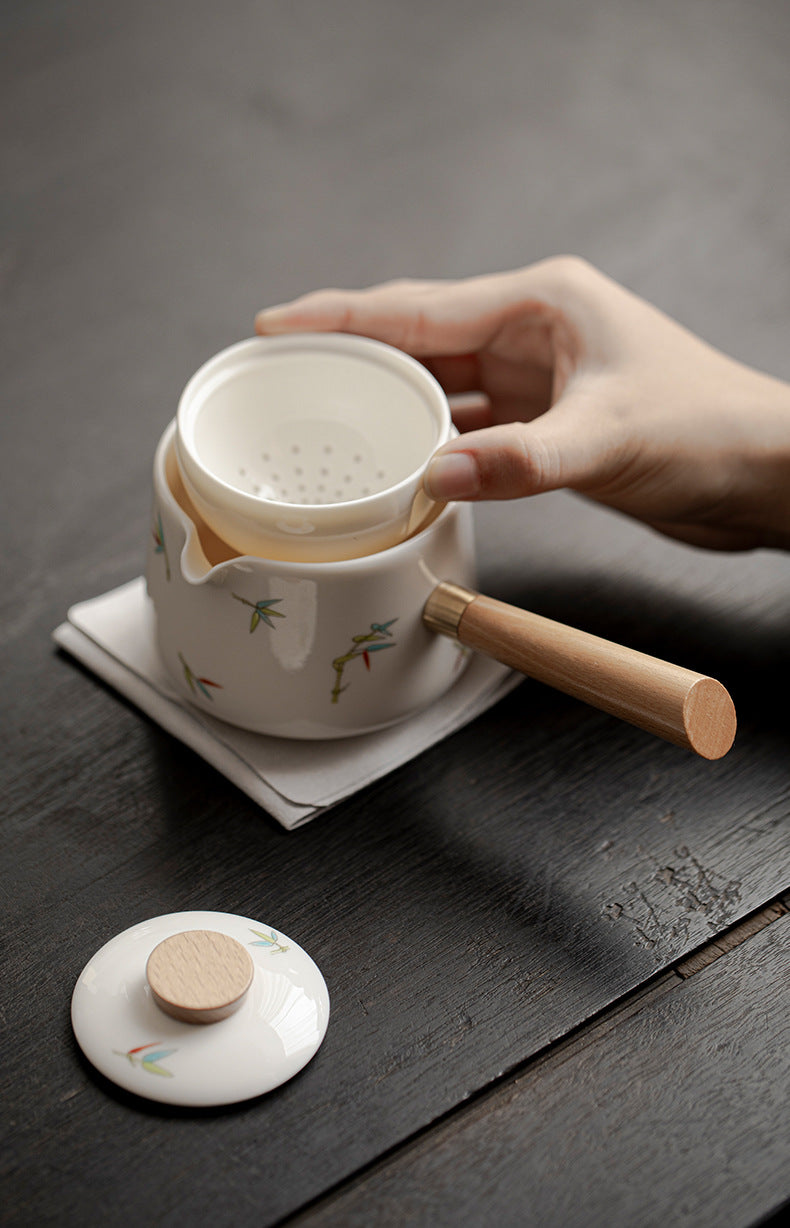 Person holding a white ceramic teapot with wooden handle on a dark wooden surface