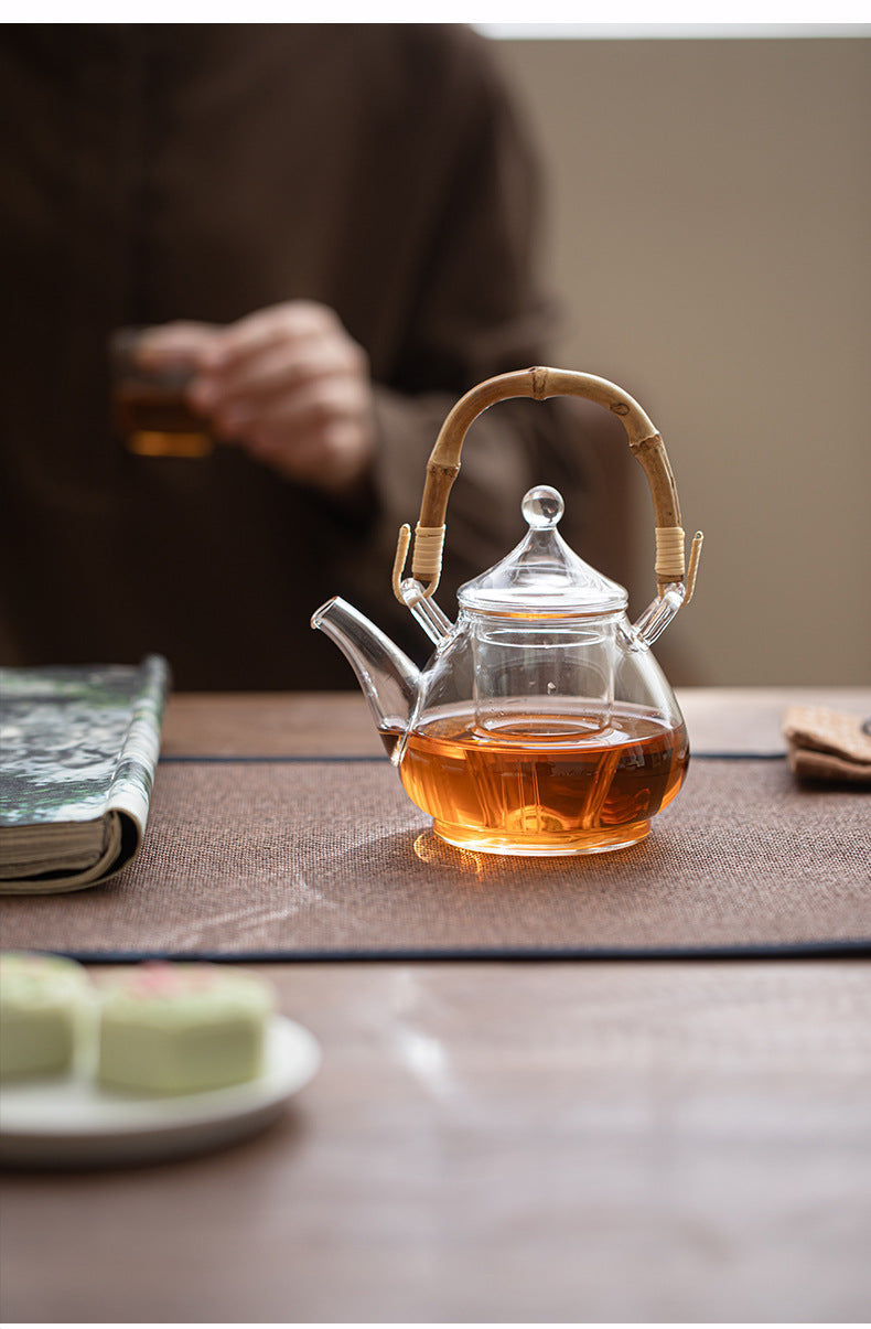 A clear glass teapot filled with amber tea, featuring a bamboo handle, set on a brown tea mat on a wooden table. A person holding a glass cup is blurred in the background.