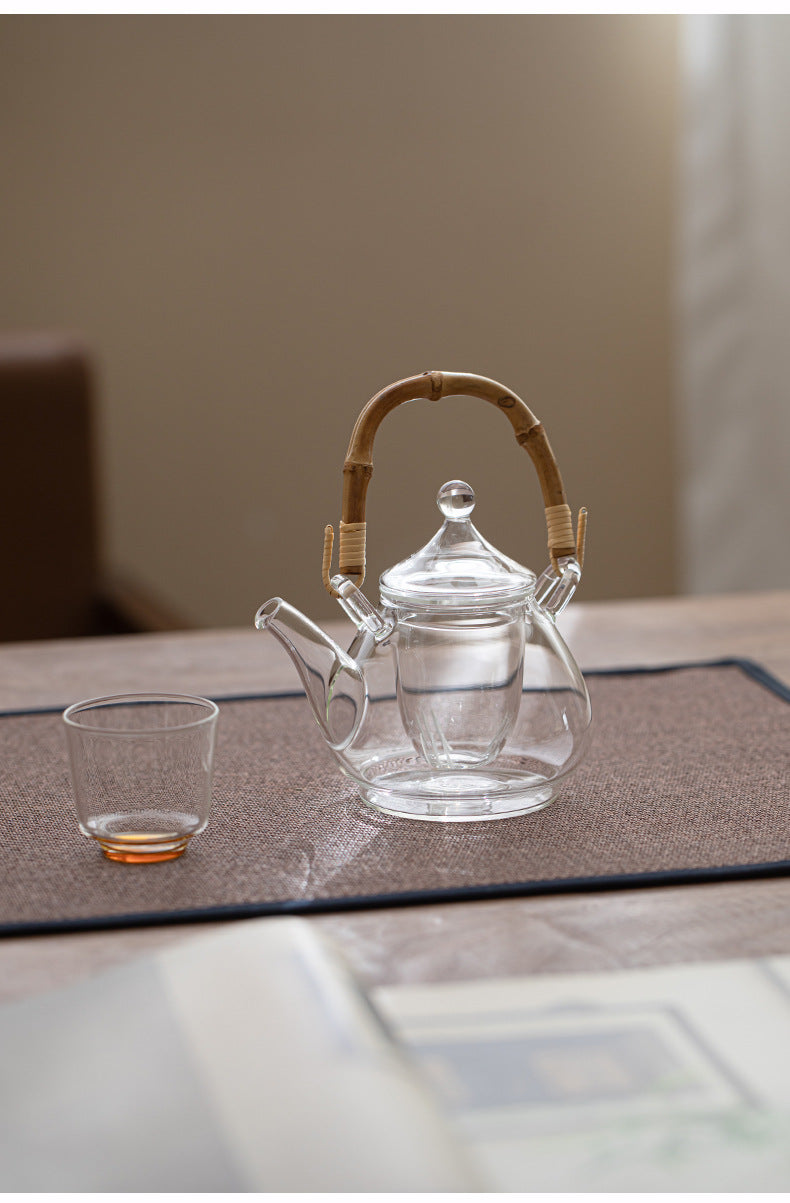 Clear glass teapot with a bamboo handle and matching glass cup on a dark placemat, set on a tabletop in a softly blurred indoor setting.