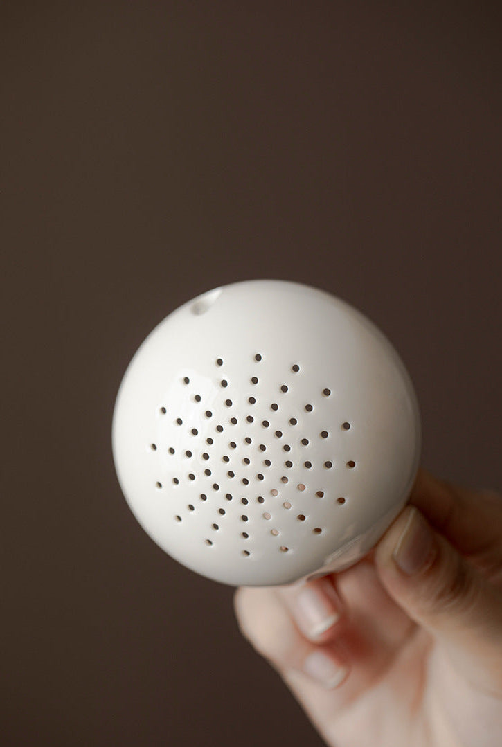 White spherical tea infuser held by a hand against a brown background