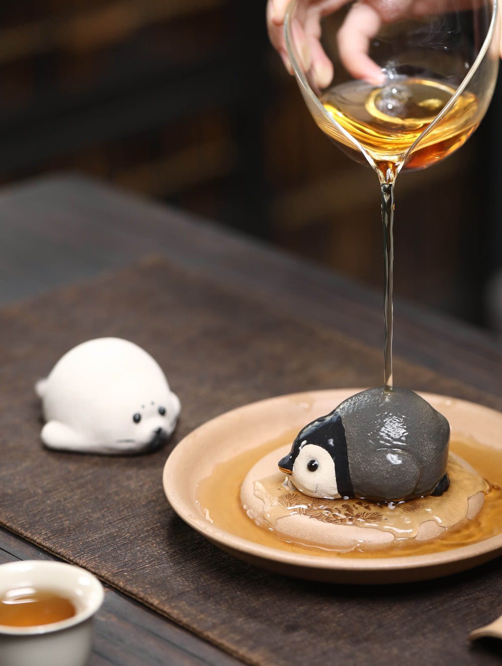 Tea poured over the penguin tea pet on a small dish, the wet clay turning darker, seal tea pet nearby