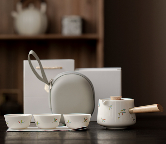 Ceramic tea set with teapot and cups on a wooden surface, featuring a carrying case in the background.