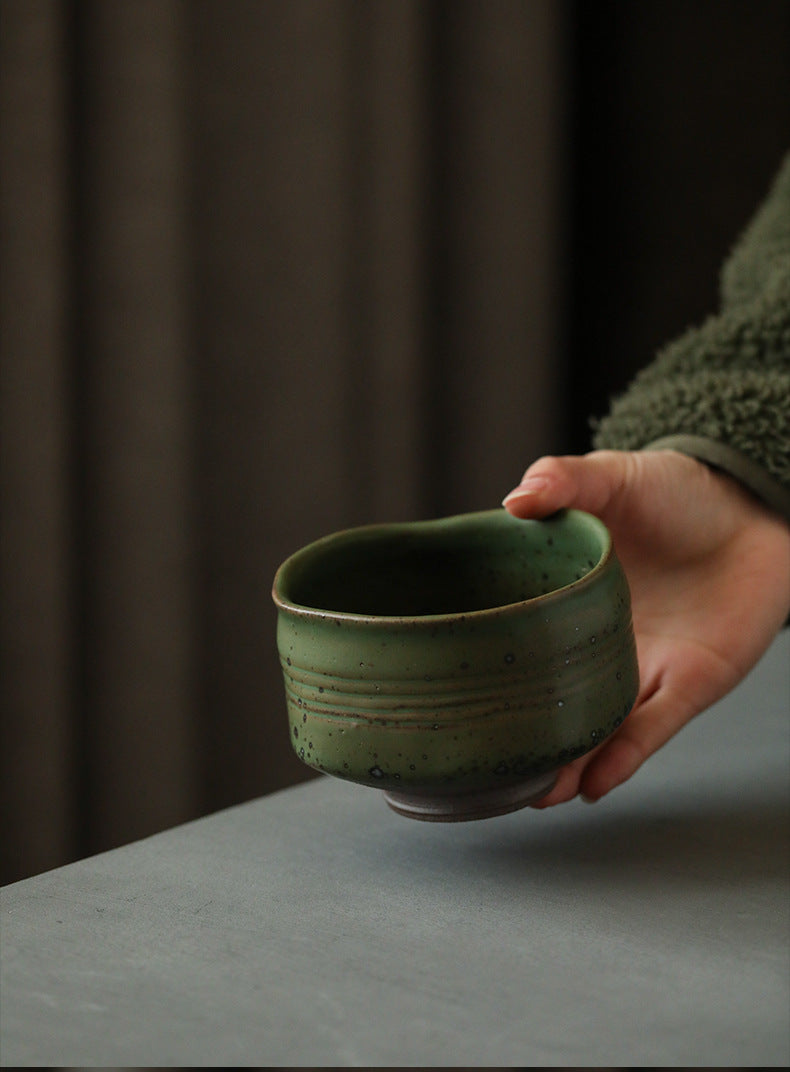 Hand holding a green ceramic Matcha bowl against a dark curtain background