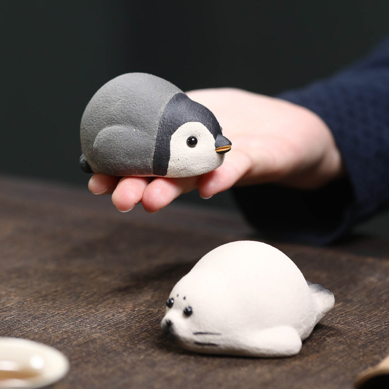 Palm-sized clay penguin tea pet held in an open hand, seal tea pet on the tea table below
