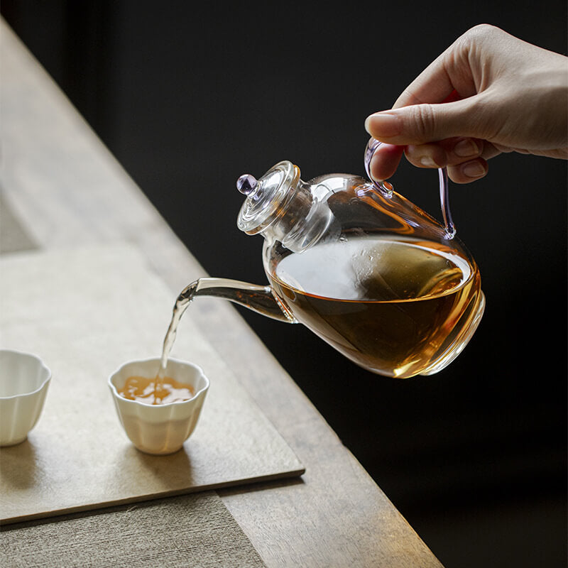 Person pouring tea from a glass teapot into a small cup on a wooden surface.