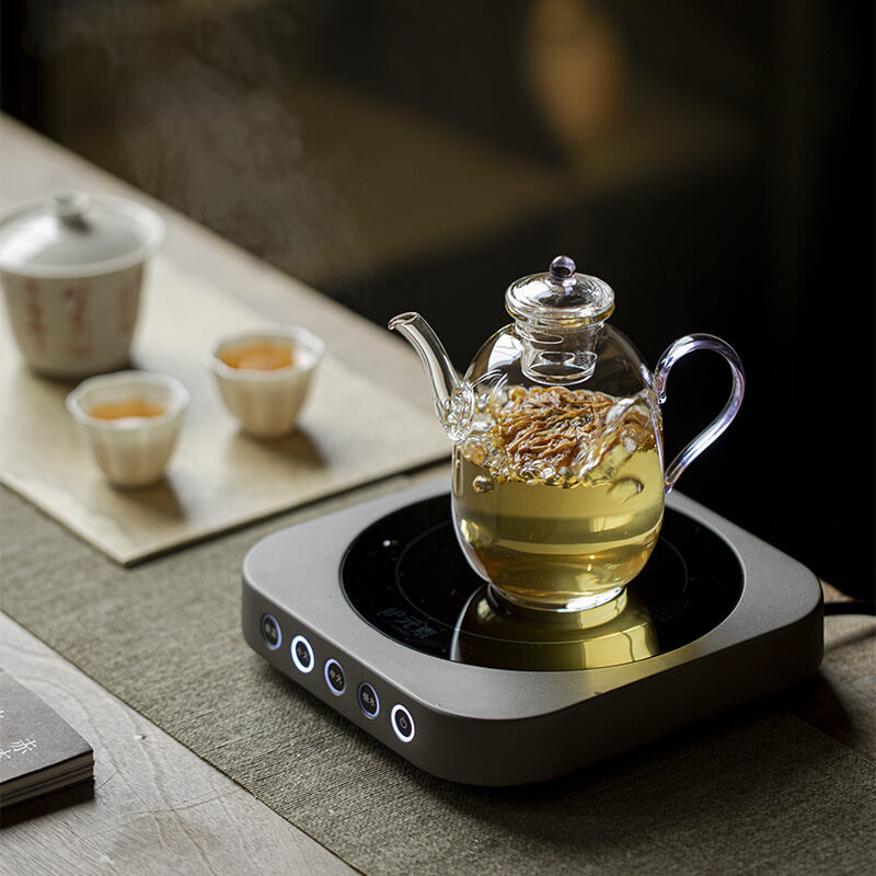 Glass teapot with tea on a heating pad on a wooden table.